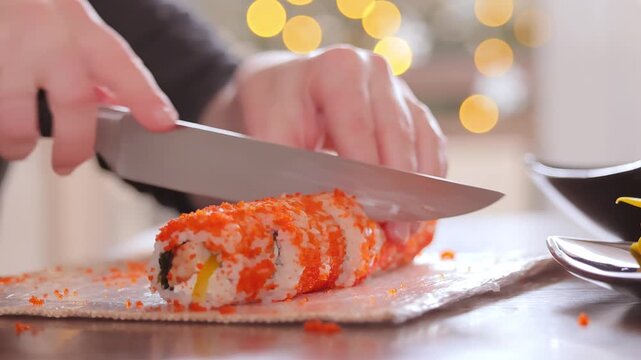 Close up of professional chef using sharp kitchen knife to slice fresh sushi roll into even pieces on wooden table.