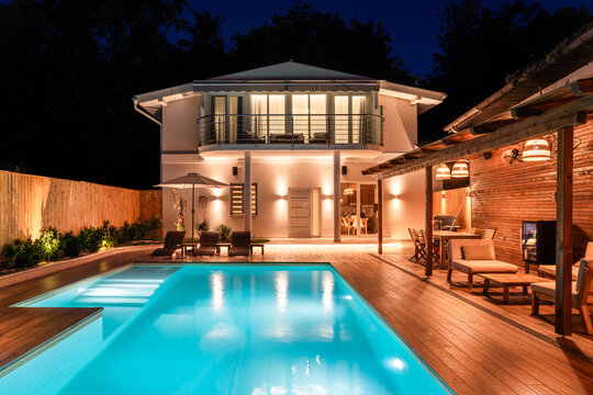 Luxury modern house with illuminated swimming pool and outdoor lounge area at night. Elegant backyard featuring wooden deck and cozy lighting
