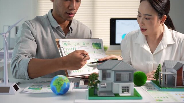 Two business people review green energy charts with house models, a wind turbine and a globe, discussing sustainable housing and eco planning in a bright office.