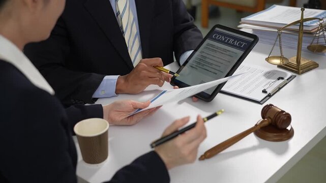Two business people review a contract on a tablet at a desk with legal papers, a gavel and balance scale, showing agreement, law and professional consultation.