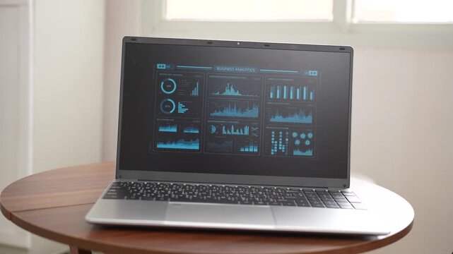 Laptop on a round wooden table showing data charts and dashboard graphs in a bright home or office space with soft natural light.