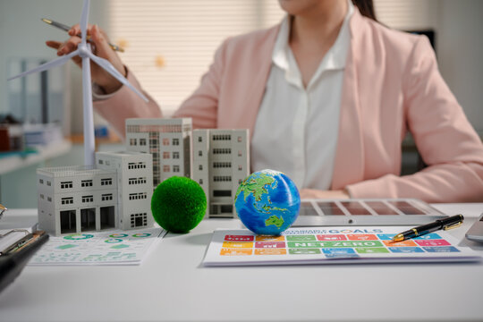 Businesswoman planning sustainable city development with wind turbine model, buildings, globe and energy chart on office desk.