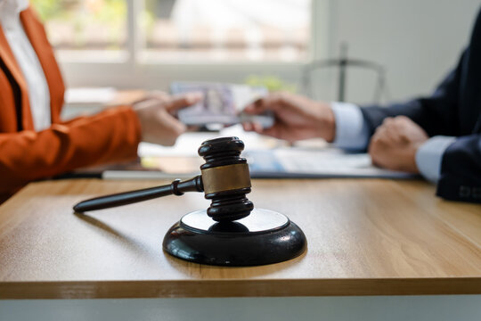 Judge gavel on a desk in focus with two people in suits exchanging money or papers in a blurred office, suggesting legal dispute, settlement, corruption, or bribery.