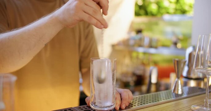 Drink making. Cold cocktail. Bartender hand stirring ice cube in tall glass with bar spoon preparing refreshing ingredient on pub counter.