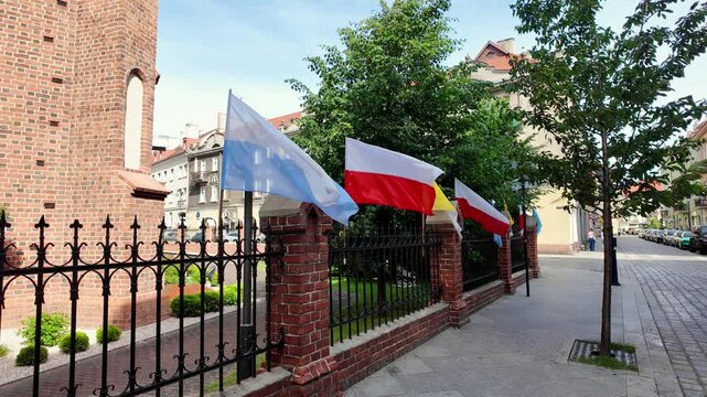 Marian, Polish, and papal flags waving on a metal fence with brick pillars next to a red brick gothic church, featuring a city street, old town houses, trees, and pedestrians in bright sunlight.