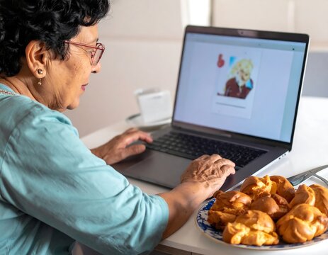 Senior woman using laptop at kitchen table with plate of pastries