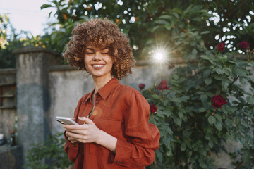 Obraz premium Joyful woman enjoys an outdoor lifestyle moment, smiling while using a smartphone in a sunny garden, wearing a rust red blouse among flowers, capturing candid warmth and charm