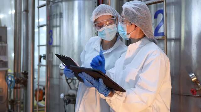 Female quality control inspectors in protective gear conducting audit in modern industrial plant, Professional food safety auditors wearing face masks and lab coats monitoring manufacturing equipment
