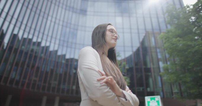A young Caucasian businesswoman or student or intern in a business suit and glasses in an urban setting among trees and modern buildings stands with her arms crossed - orbital shot