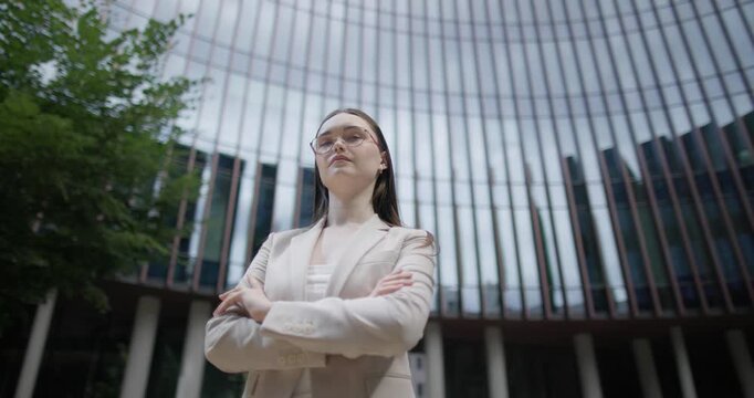 A young Caucasian businesswoman or student or intern in a business suit and glasses in an urban setting among trees and modern buildings stands with her arms crossed - orbital shot