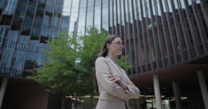 A young Caucasian businesswoman or student or intern in a business suit and glasses in an urban setting among trees and modern buildings stands with her arms crossed - orbital shot