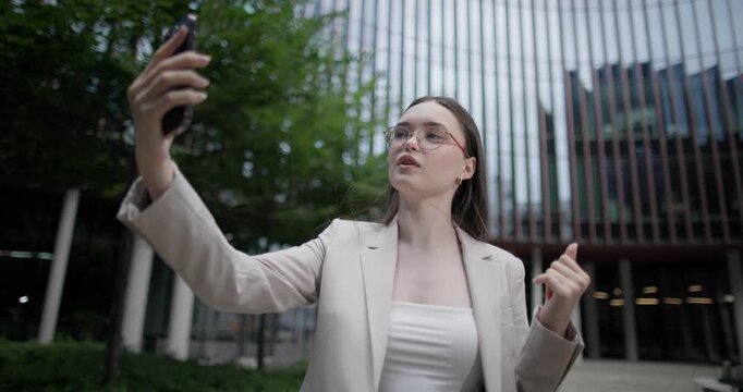 Young Caucasian businesswoman or student or intern in a business suit and glasses in an urban setting among trees and modern buildings making a video call on the phone