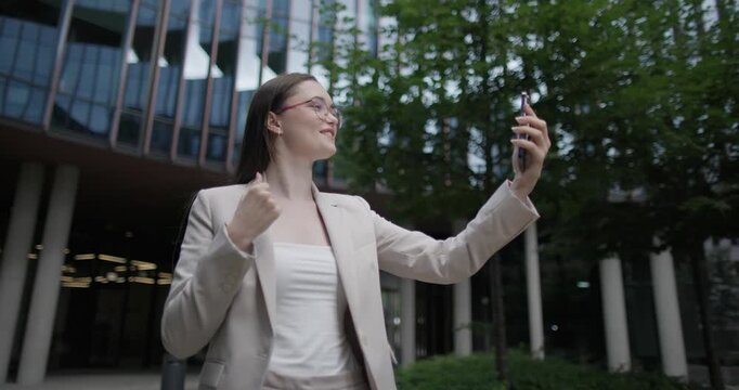 Young Caucasian businesswoman or student or intern in a business suit and glasses in an urban setting among trees and modern buildings making a video call on the phone
