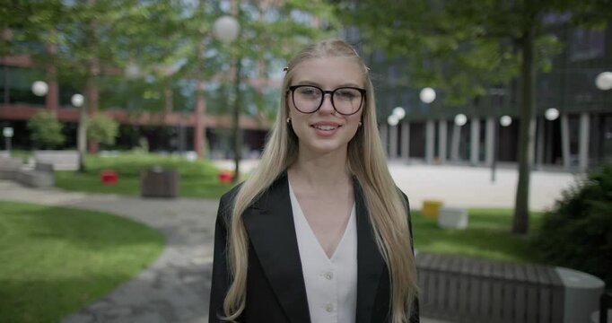 A young Caucasian woman who studies daily wearing glasses stands in an urban setting among houses and trees and looks at the camera.