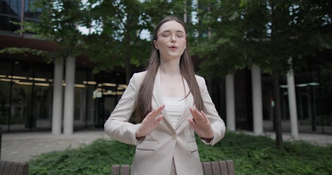 A young Caucasian businesswoman or student or intern in a business suit and glasses in an urban setting among trees and modern buildings does breathing exercises and meditates to relieve stress 