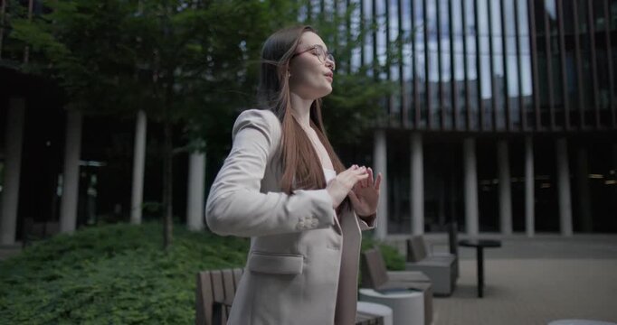 A young Caucasian businesswoman or student or intern in a business suit and glasses in an urban setting among trees and modern buildings does breathing exercises and meditates to relieve stress 