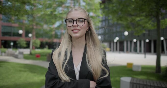 A young Caucasian woman who studies daily wearing glasses stands in an urban setting among houses and trees, crosses her arms over her chest and looks at the camera.