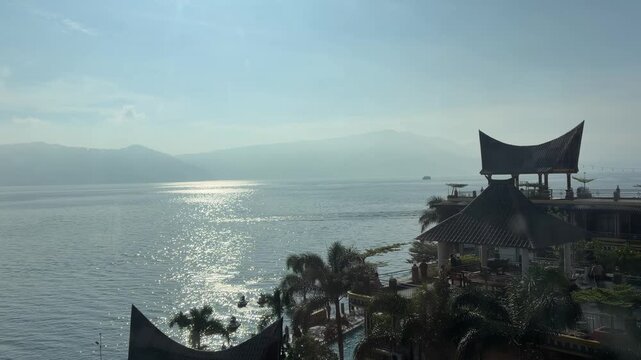 Panoramic of a clear sunrise at Lake Toba with a traditional Batak style resort terrace in the foreground, bright sunrays cutting through clouds