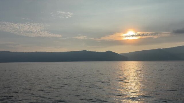 Panoramic time-lapse of a sunset at Lake Toba with a traditional Batak style resort terrace in the foreground, bright sunrays cutting through clouds