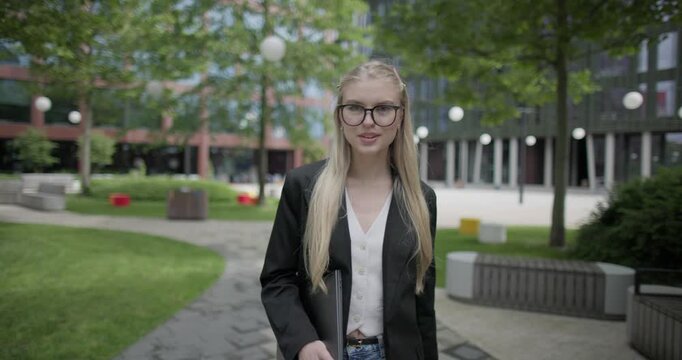 A young Caucasian woman who studies daily with glasses and a laptop in a modern urban environment among houses and trees walks through a park
