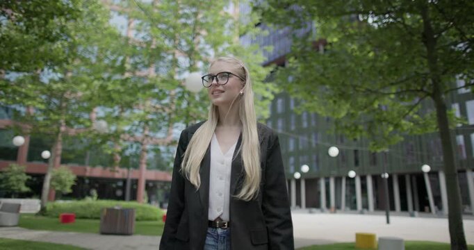 A young Caucasian woman who studies daily wearing glasses in a modern urban environment among houses and trees walks through a park