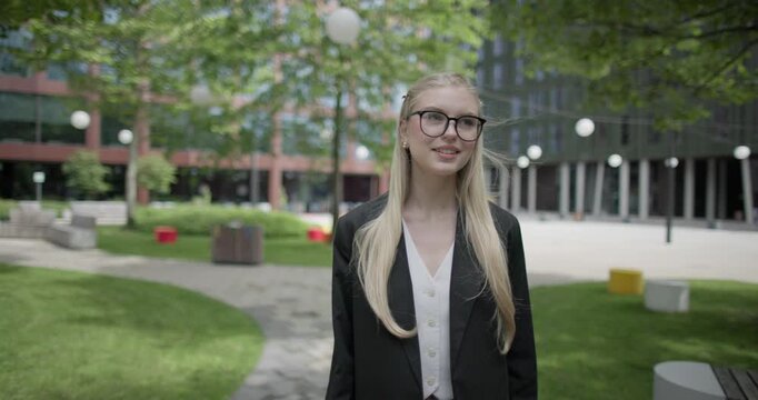 A young Caucasian woman who studies daily wearing glasses in a modern urban environment among houses and trees walks through a park