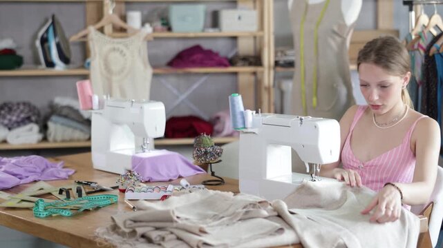 Skillful young tailoress sewing textile sitting at her workplace in tailor shop