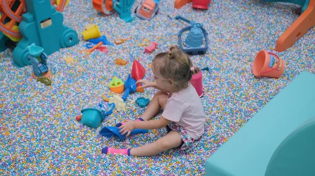 Little toddler girl using a toy spade and bucket to scoop colorful plastic balls in an indoor sandbox play area. Childhood and sensory play concept