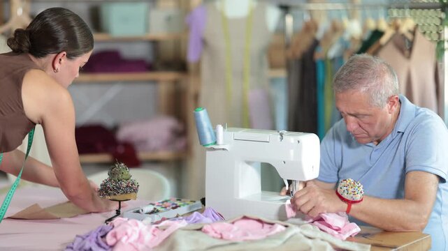 Working day in sewing workshop, textile industry. Mature man master sew clothes in sewing machine, girl pins paper pattern to fabric, attaches template to fabric, preparing to cut fabric into pieces. 