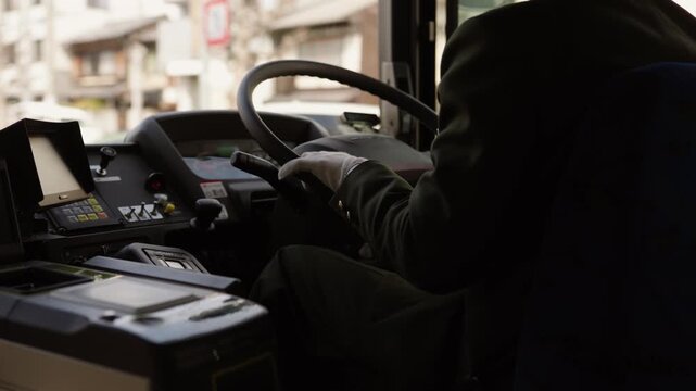 Japanese bus driver in uniform and white gloves gripping steering wheel at driver console in urban Japan.