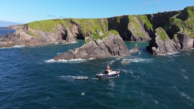 A boat is anchored near rugged cliffs and rocky outcrops at Dunquin Pier. The vibrant green landscape contrasts with the deep blue sea. Great for travel or nature content.
