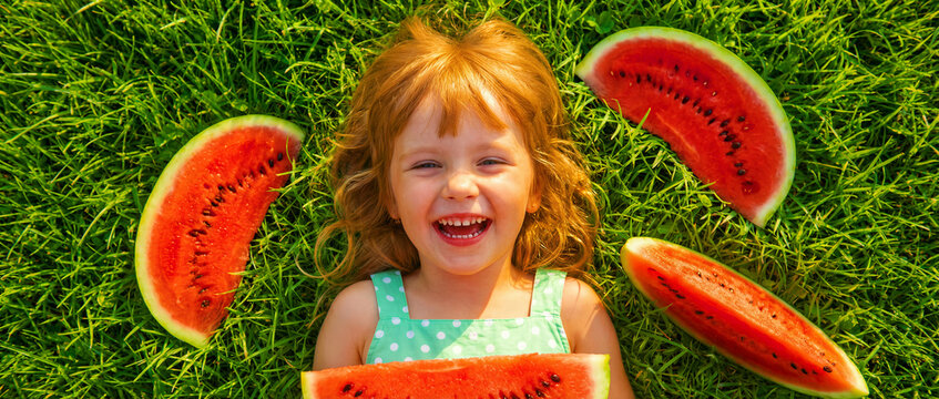 Happy redheaded girl lying on green grass with watermelon slices. Child laughing and enjoying fresh fruit during summer. Healthy lifestyle concept, ideal for family and nutrition marketing.