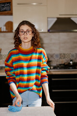 Obraz premium A young woman in a colorful striped sweater stands at a kitchen counter, glove on one hand, looking up with a surprised expression. A candid home lifestyle moment.