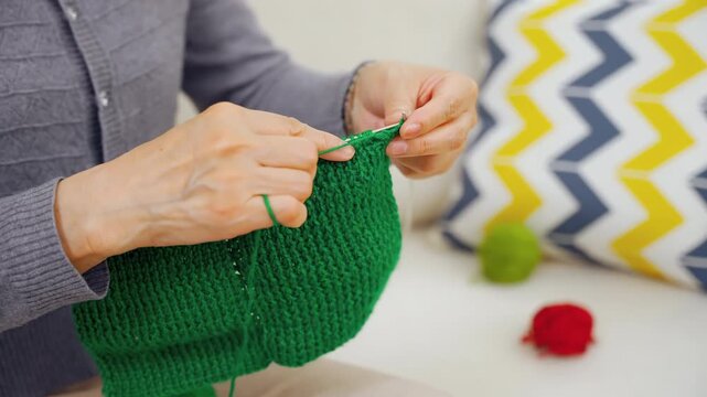 Talented elderly grandmother woman knitting green sweater by yarn ball and needles with wrinkly hands crafting hobby in free time sitting comfortably on sofa. People, age and leisure.