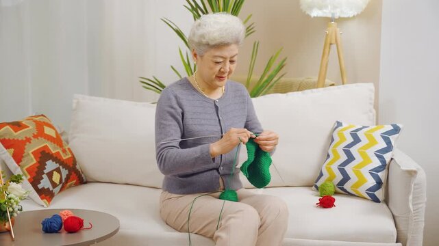 Focused elderly woman knitting sweater by yarn ball and needles with wrinkly hands crafting hobby in free time sitting comfortably on sofa. People, age and leisure.