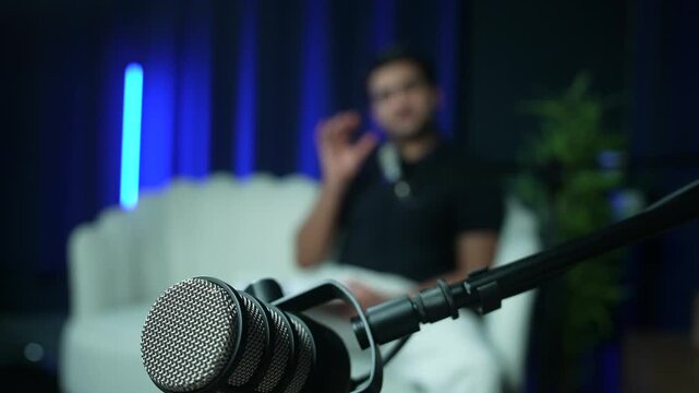 Podcast host speaking beside microphone in blue lit recording studio, blurred with stand opposite in focus