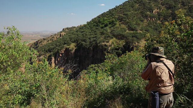 Photographer with camera equipment standing among vegetation while overlooking rugged canyon cliffs and mountain terrain near La Garita Canyon, Sierra P&eacute;njamo, Guanajuato, Mexico.
