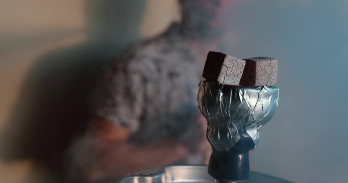 smoking shisha in the background while hot charcoal cubes sit on a foil-covered hookah bowl. Suitable for hookah lounge, tobacco smoking, relaxation, nightlife, leisure, and social lifestyle concepts.