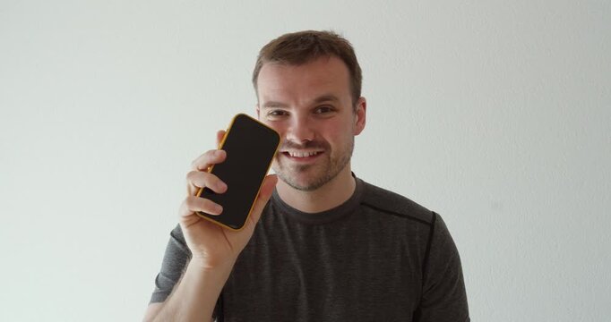 smiling man holding a smartphone toward the camera and pointing at the display. Suitable for mobile apps, advertising, technology, communication, marketing, promotions, and digital concepts.