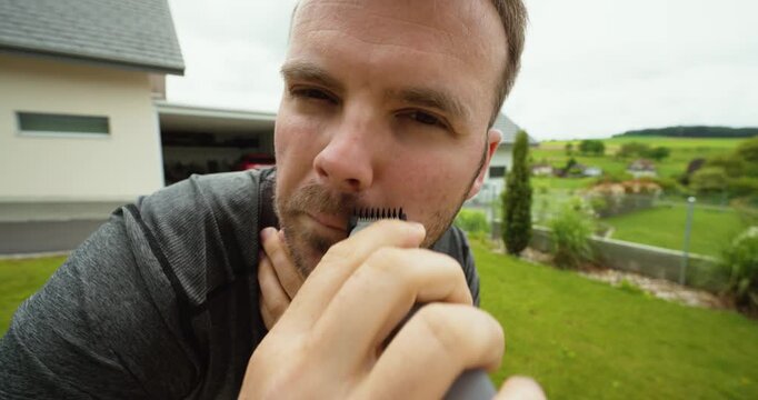 Extreme close-up stock video showing a man trimming beard hair with an electric shaver outdoors in a garden environment. Suitable for grooming, beard care, men&rsquo;s hygiene, self-care routines