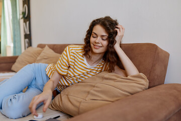 Obraz premium Relaxed woman lounging on a beige sofa at home, wearing striped tee and blue jeans. Casual lifestyle setting emphasizes comfort, relaxation, and everyday moments. Soft natural light highlights a cozy