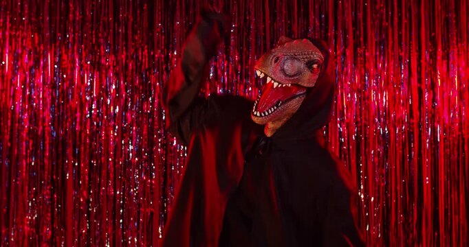 Halloween funny dance of scary dinosaur in black costume and dragon mask on fun party. Creepy monster performance with teeth, cape and skeleton gloves dancing under red lighting by tinsel curtains