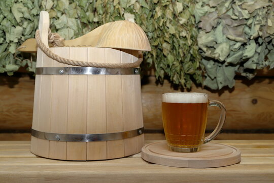 A mug of light beer on a circle wooden tray and a wooden bucket with a wooden ladle are on a plank bench against the background of a log wall with a set of dry birch and oak brooms is hanging on it.