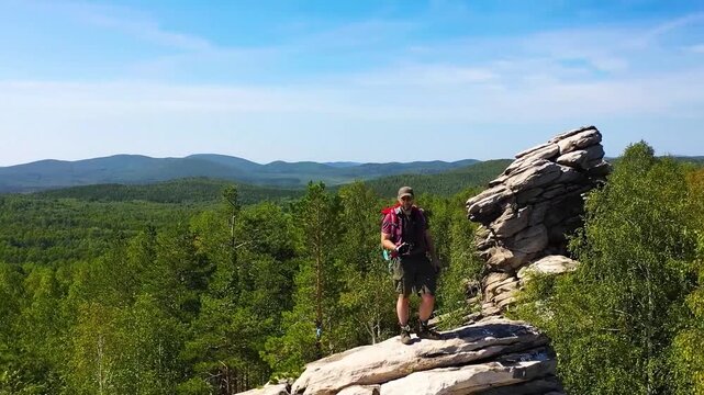 The camera pans around a middle-aged Caucasian male tourist with a large backpack and a camera in his hands. Standing on a rocky mountain in the middle of a forest, he surveys the landscape.