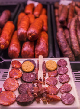 Primer plano de un plato con rodajas de chorizo, salchich&oacute;n, jam&oacute;n y galletas saladas para clientes. Al fondo, embutidos tradicionales expuestos en el mostrador de una charcuter&iacute;a.