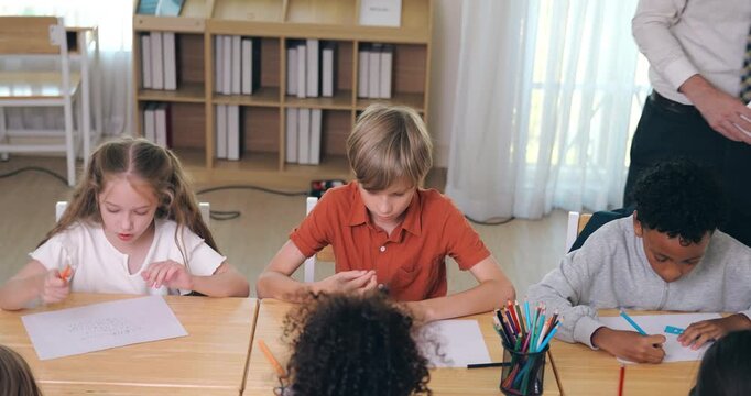 A caring male teacher walking around and supervising a diverse group of students writing and drawing at their desks.