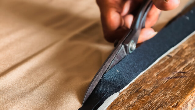 Close-up of a craftsman's hand cutting black leather material with scissors on a wooden workbench