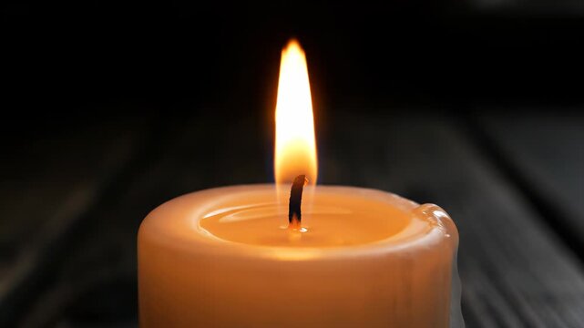 Close-up of single thick white pillar candle burning with steady yellow flame in dark environment. Detailed macro shot of glowing wick, melted wax pool, and flickering fire on wooden surface.