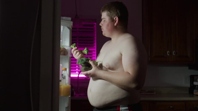 Overweight man opens fridge at night. Overweight guy eats broccoli. Healthy overweight lifestyle. Night eating changes concept. Diet man eats broccoli at night. Diet healthy weight loss.