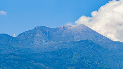 Obraz premium Majestic volcanic mountain peak rising under a clear blue sky with soft white clouds, showcasing the breathtaking grandeur and untouched beauty of nature's powerful landscapes
